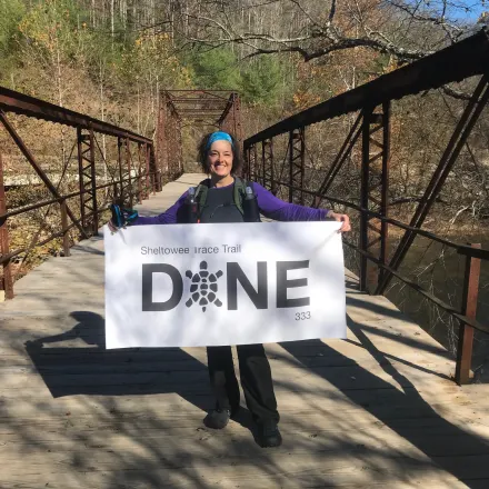 Dr. Schlegel's staff photo from College Mall Veterinary Hospital where she is holding up a sign on a lake bridge that says Sheltowee Irace Trail Done 333. 